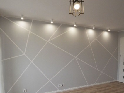 Szara ściana z białymi geometrycznymi wzorami, oświetlona punktowymi lampami sufitowymi i designerską lampą wiszącą.