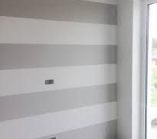 Ściana pomalowana w poziome pasy w odcieniach szarości i bieli, widoczne gniazdka elektryczne, kadr z fragmentem okna.