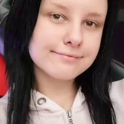 Portret młodej kobiety z długimi czarnymi włosami w jasnej bluzie z kapturem, siedzącej na czarno-czerwonym fotelu gamingowym.