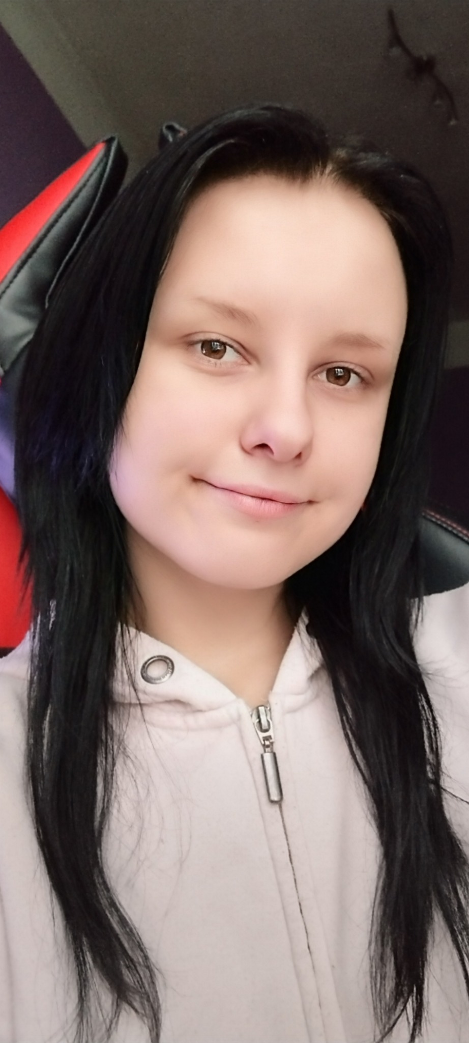 Portret młodej kobiety z długimi czarnymi włosami w jasnej bluzie z kapturem, siedzącej na czarno-czerwonym fotelu gamingowym.