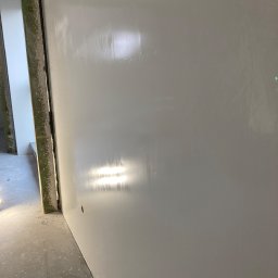 Sergiej Krykun - Świeżo wykończona, gładka ściana w jasnym odcieniu szarości, z widocznym otworem na gniazdko elektryczne i fragmentem ościeżnicy w tle, ukazująca efekt równomiernie nałożonej warstwy wykończeniowej.