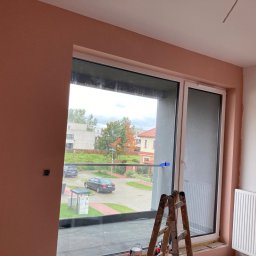 Sergiej Krykun - Pokój w trakcie wykańczania, z częściowo nałożoną gładzią w odcieniu brzoskwiniowym, widoczne okno z widokiem na ulicę i drabina malarska.