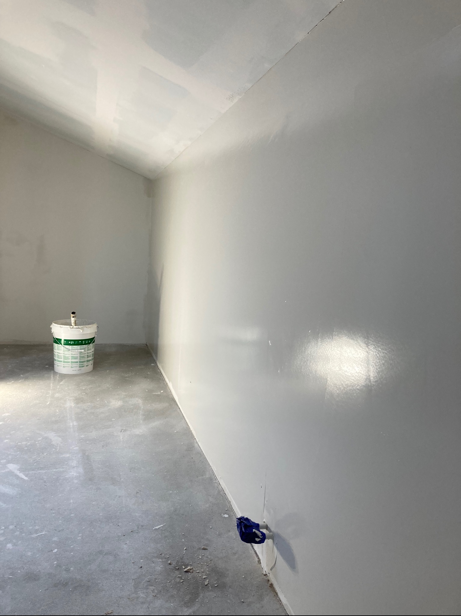 Świeżo pomalowane na szaro pomieszczenie ze śladami gładzi na suficie, wiadro z farbą w rogu i zabezpieczenie folią przy podłodze.