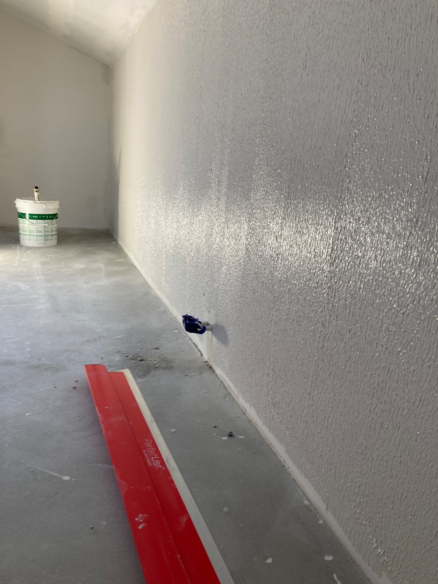 Świeżo nałożona, błyszcząca gładź na ścianie w rogu pomieszczenia, widoczna puszka z farbą i czerwona szpachla Parfait Liss na podłodze.