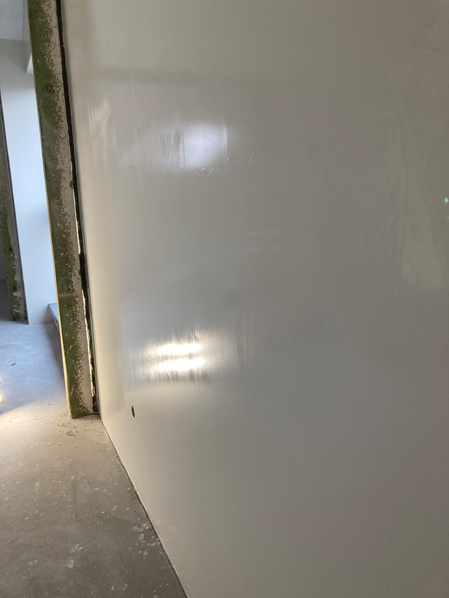 Świeżo wykończona, gładka ściana w jasnym odcieniu szarości, z widocznym otworem na gniazdko elektryczne i fragmentem ościeżnicy w tle, ukazująca efekt równomiernie nałożonej warstwy wykończeniowej.