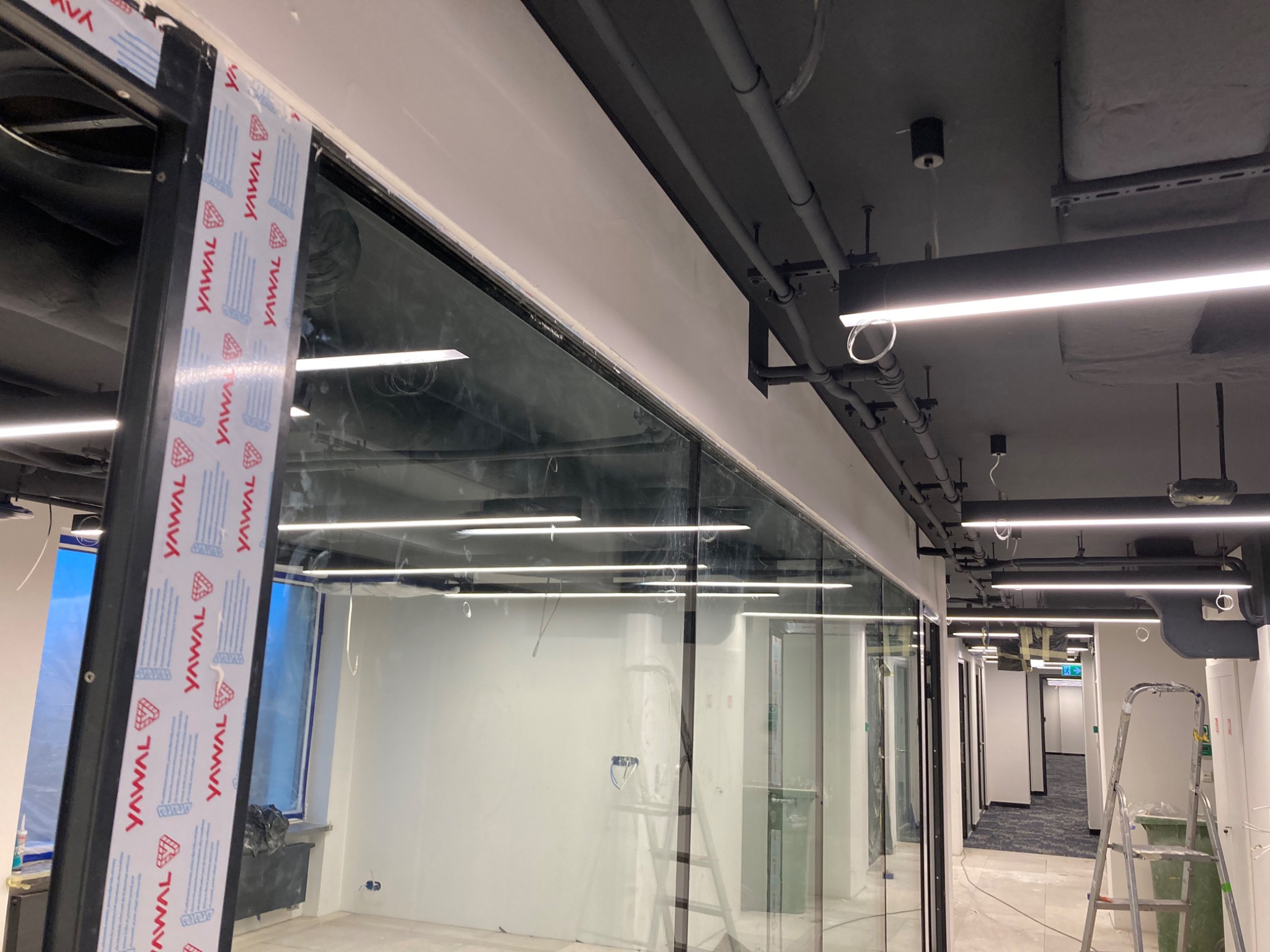 Wnętrze biurowe w trakcie wykończenia, widoczne szklane ścianki działowe z okleiną zabezpieczającą ramy, sufit z instalacjami i oświetleniem liniowym, drabina malarska i widok na korytarz.