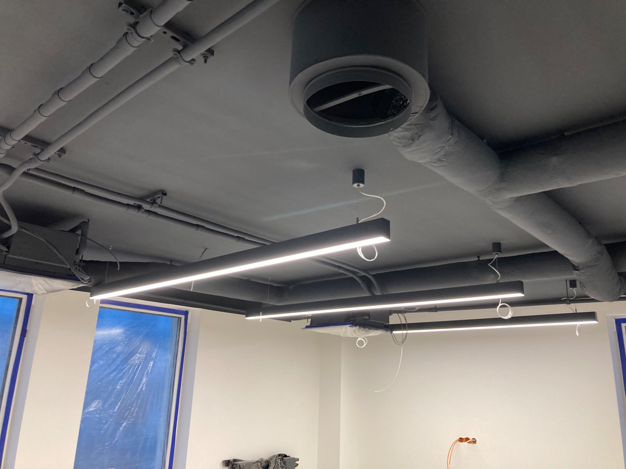 Pomieszczenie biurowe w trakcie remontu: pomalowany na szaro sufit z widocznymi rurami i wentylacją, okna zabezpieczone folią malarską, nowoczesne oświetlenie liniowe.