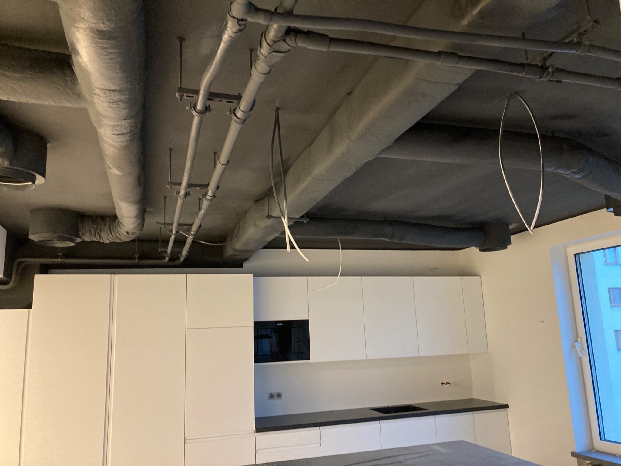 Szare rury wentylacyjne pod sufitem w surowym stanie nad białą zabudową kuchenną z czarnym blatem, widoczne wystające kable.