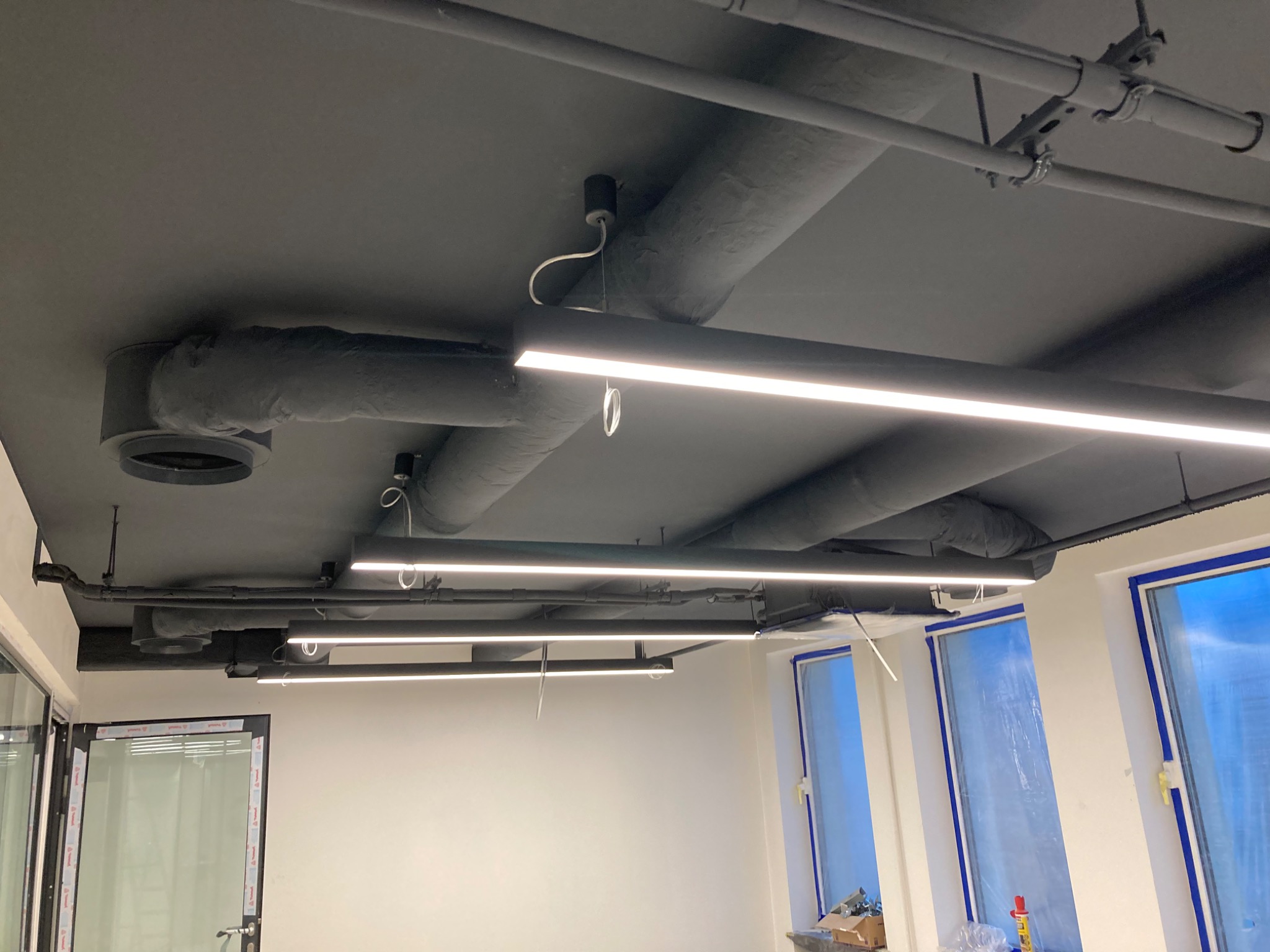 Pomieszczenie biurowe z pomalowanym na szaro sufitem, widoczne rury instalacyjne, wentylacja i oświetlenie liniowe LED. Okna zabezpieczone folią malarską.
