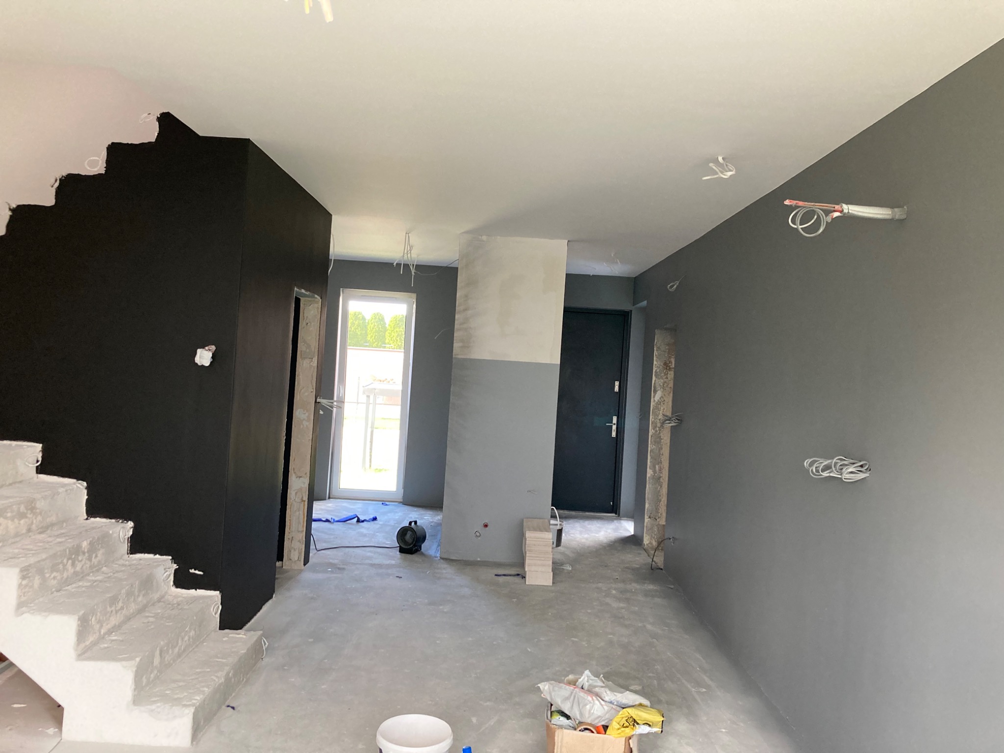 Wnętrze domu w trakcie remontu, z pomalowanymi na szaro ścianami, surowymi schodami pokrytymi gładzią i widocznymi instalacjami elektrycznymi.