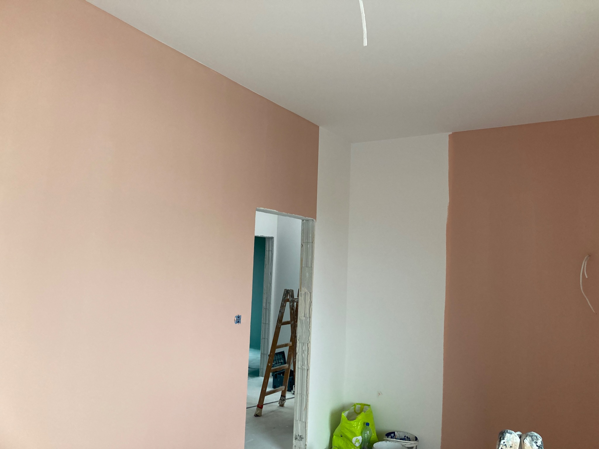 Pokój po częściowym pomalowaniu na łososiowy kolor, widoczna drabina i akcesoria malarskie, ściana z fragmentem białej farby.