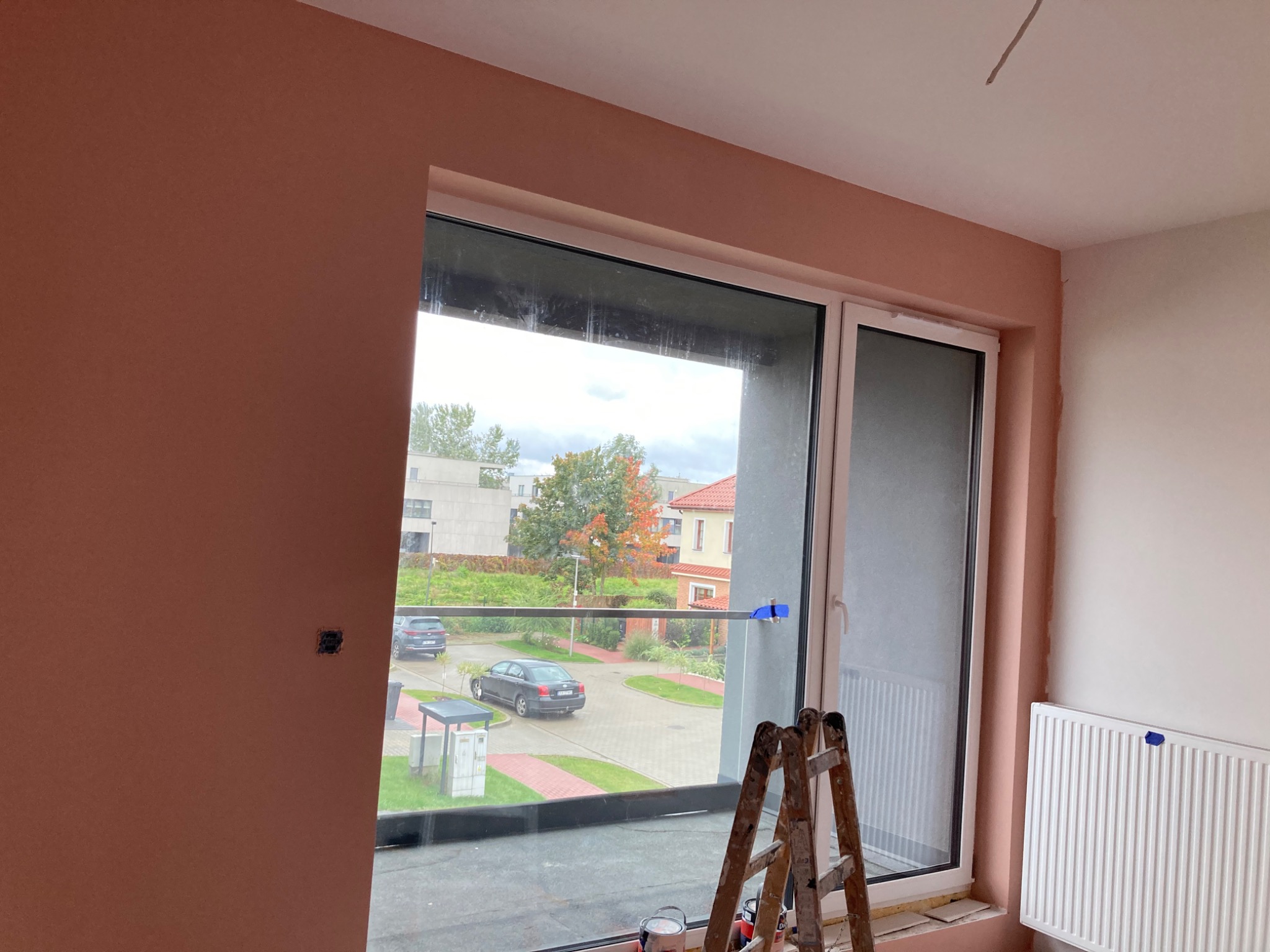 Pokój w trakcie wykańczania, z częściowo nałożoną gładzią w odcieniu brzoskwiniowym, widoczne okno z widokiem na ulicę i drabina malarska.