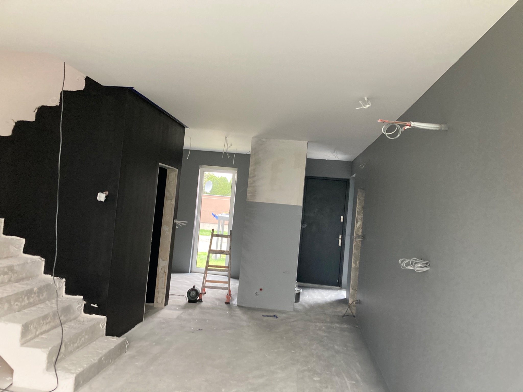 Wnętrze w trakcie malowania: szare ściany, czarna ściana przy schodach, widoczne przewody, drabina i wentylator pod oknem.