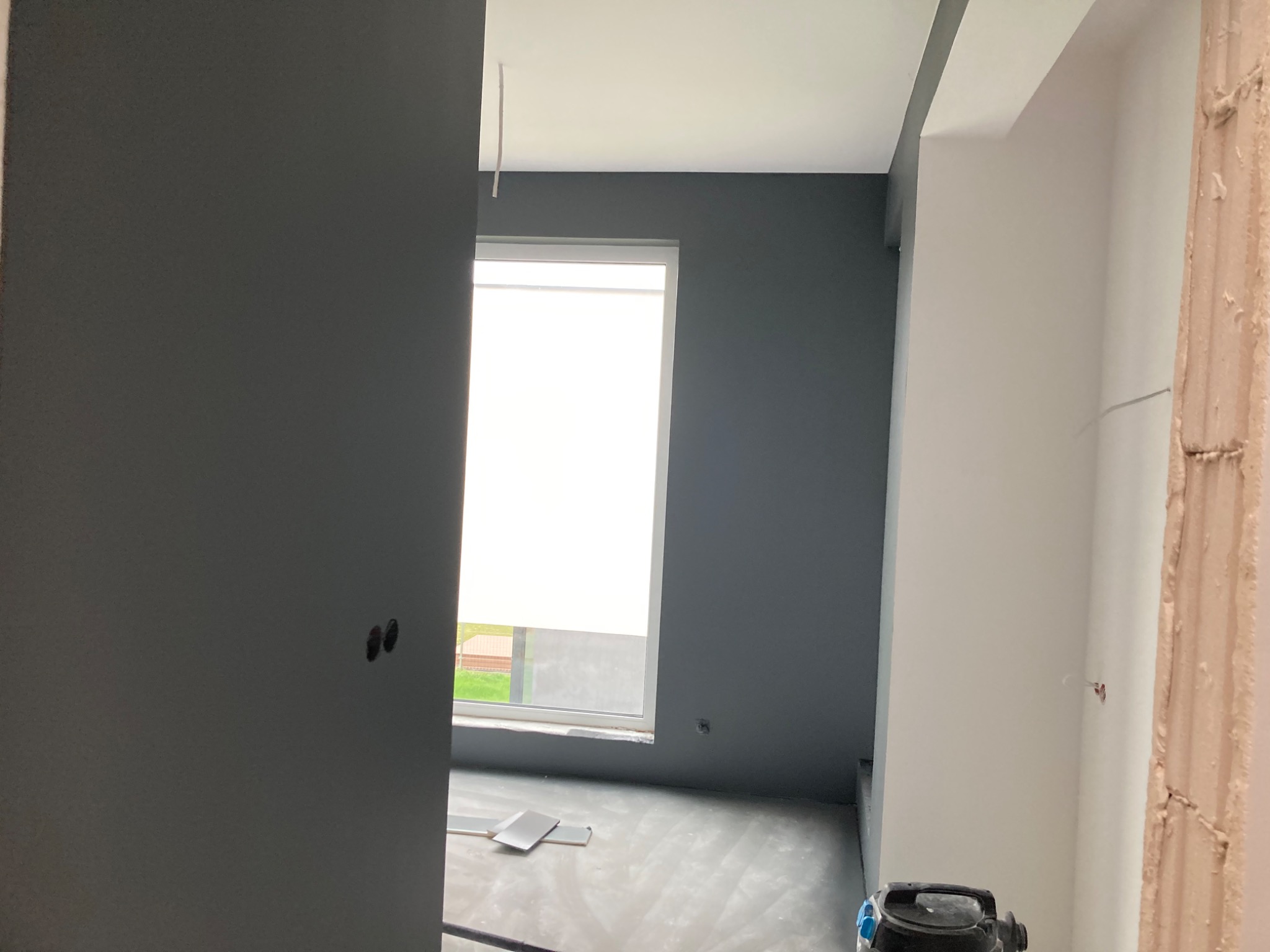 Pokój po częściowym odmalowaniu na ciemnoszaro, widoczne ślady po gładzi na podłodze i niedokończone obramowanie drzwi.