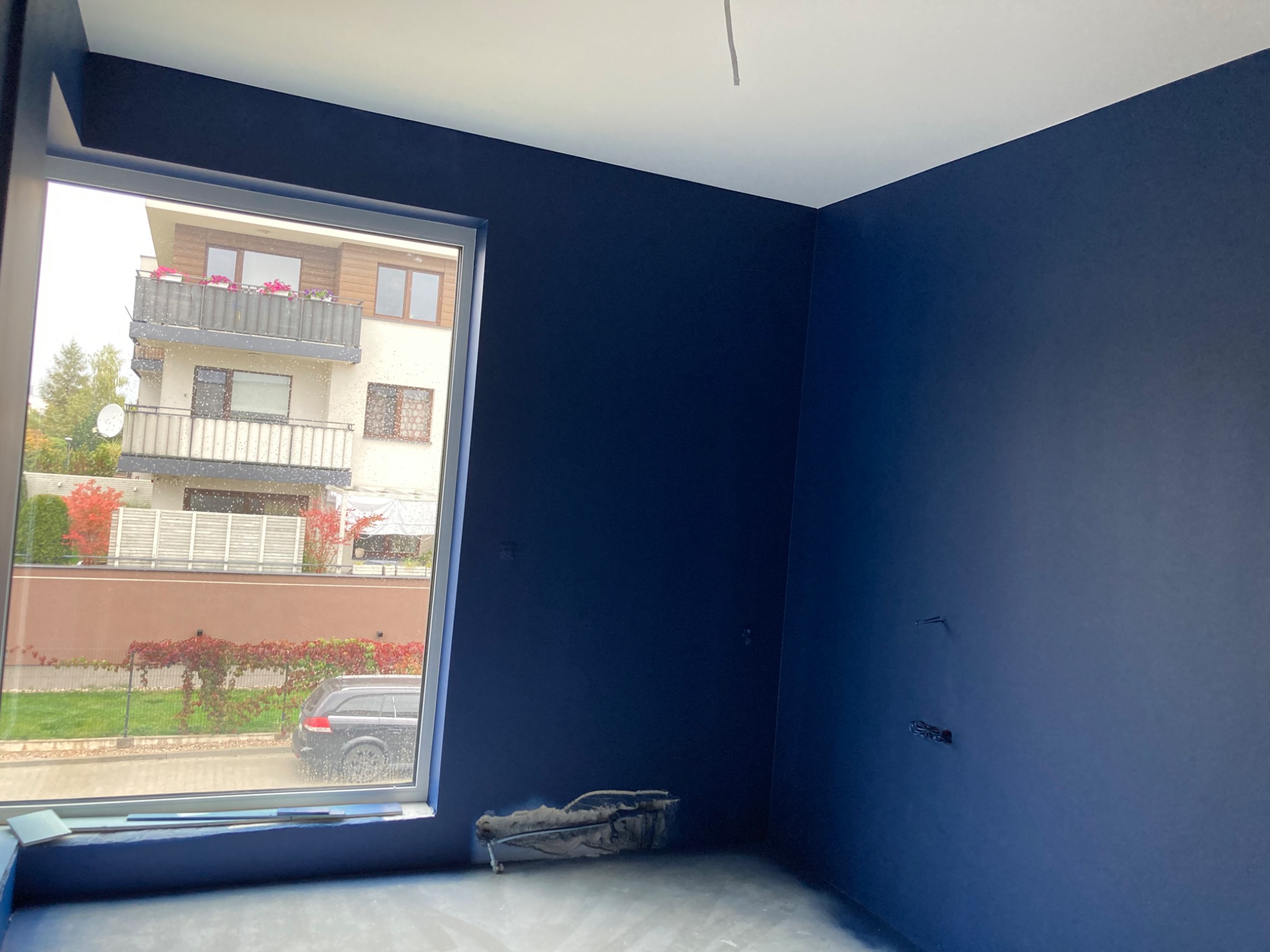 Wnętrze pokoju po malowaniu na ciemny granat, widoczne ślady po gniazdkach elektrycznych i narzędzia malarskie przy ścianie, okno z widokiem na dom i ogród.