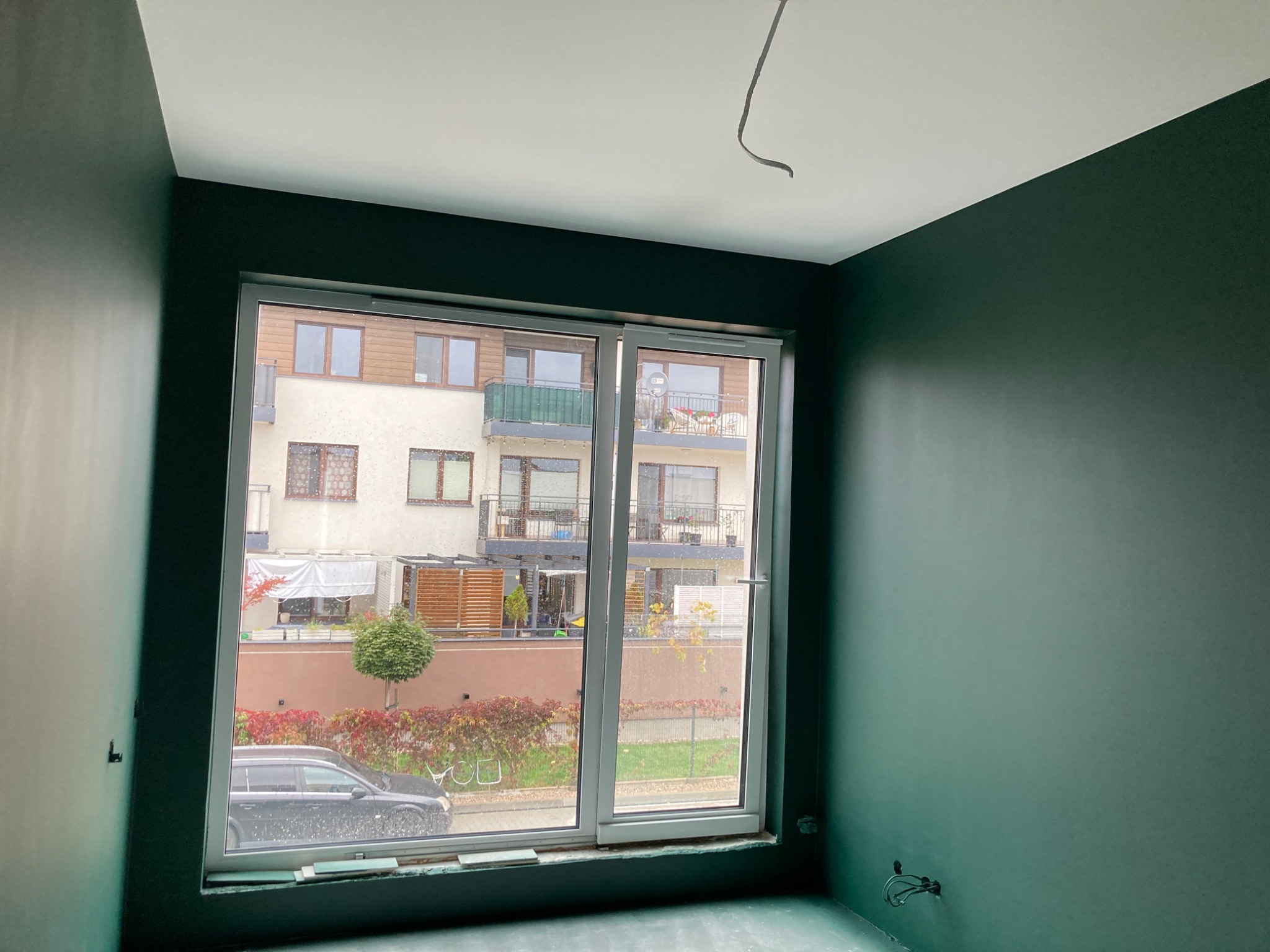 Pokój po malowaniu ścian na ciemnozielony kolor, widok na budynek mieszkalny przez okno.