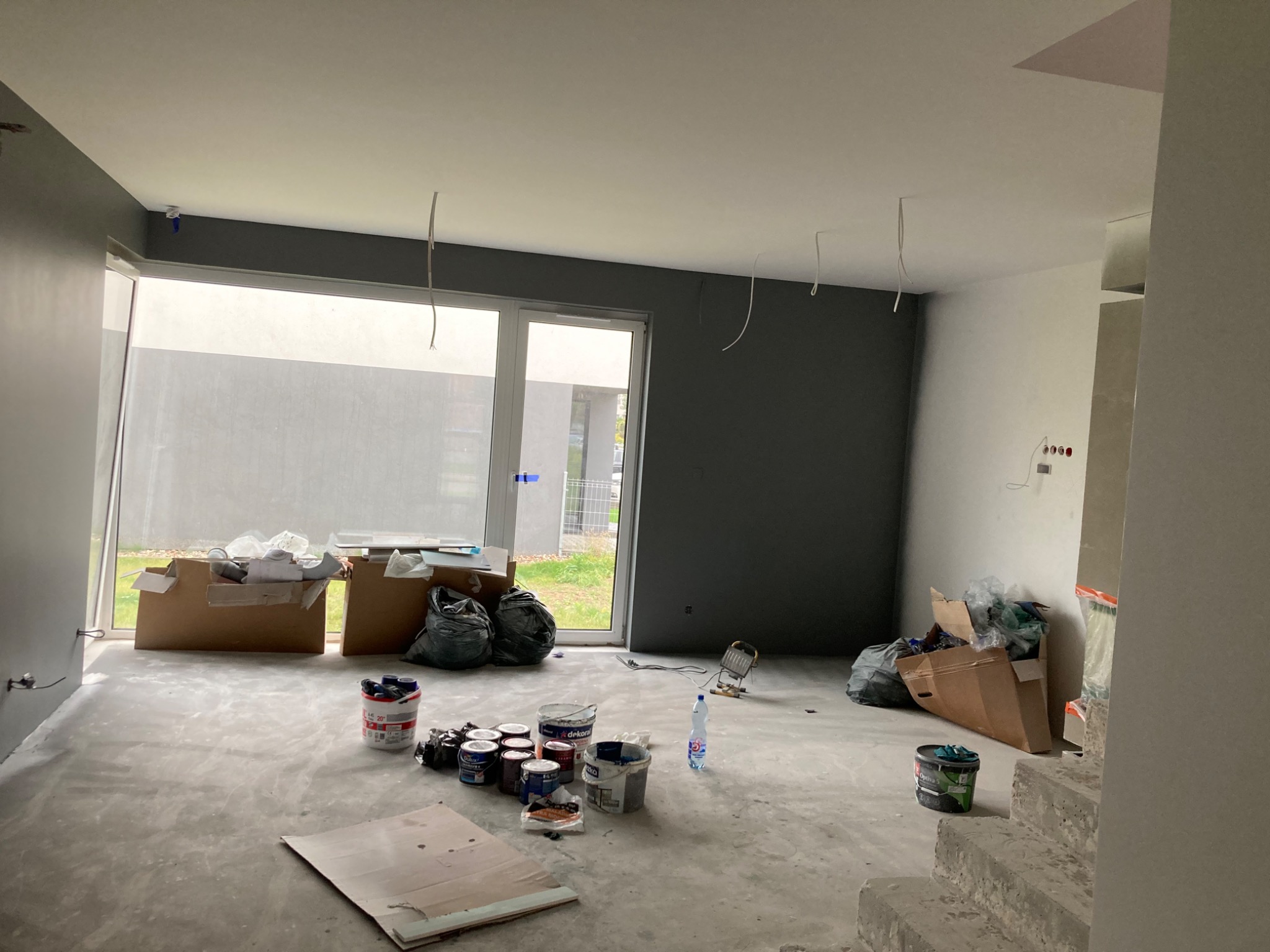 Wnętrze w trakcie remontu: szare ściany, biały sufit, puszki z farbą, narzędzia i materiały budowlane rozłożone na betonowej podłodze, widoczne kable wystające z sufitu, duże okno z widokiem na ogród.
