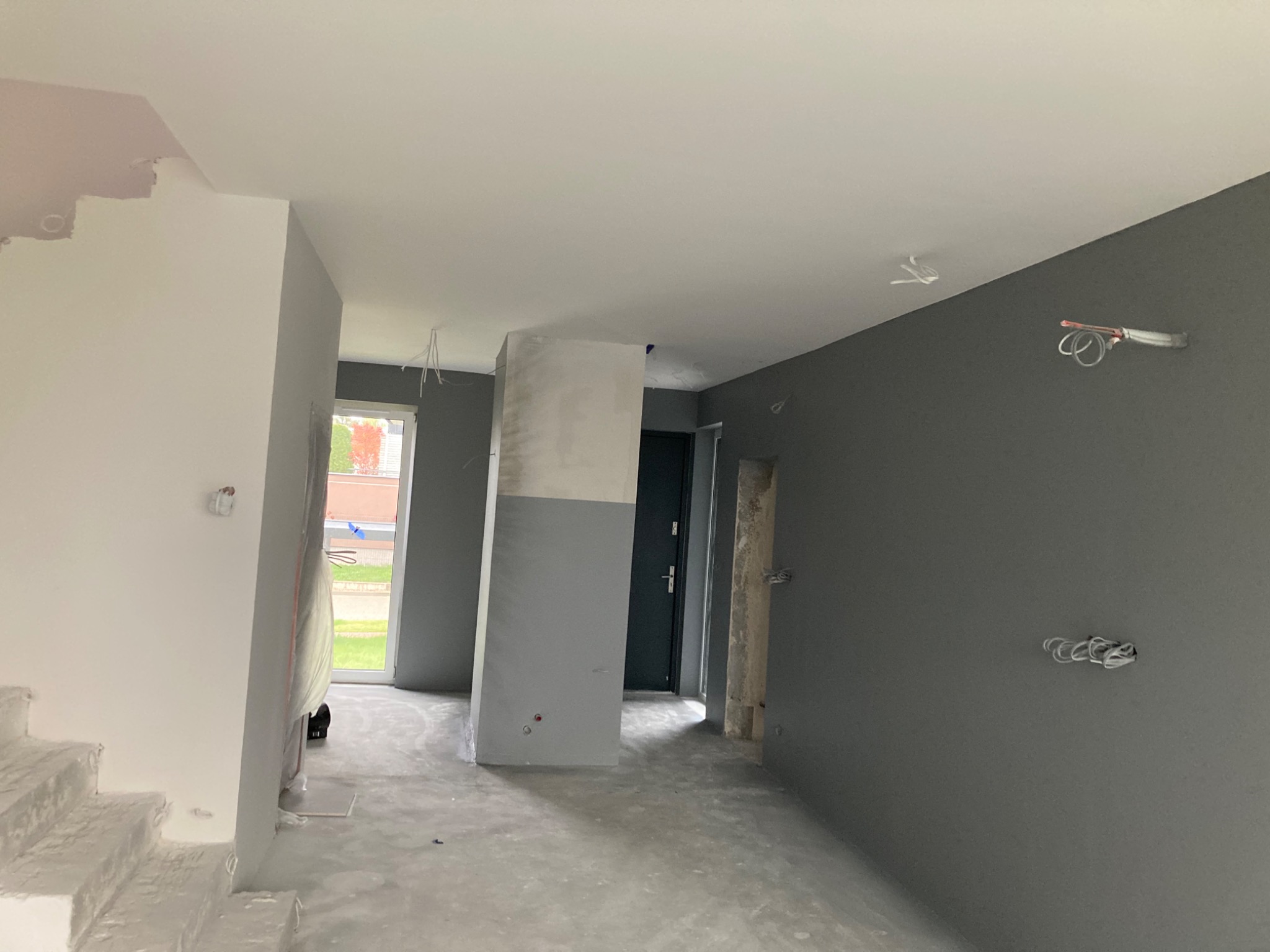 Wnętrze w trakcie prac wykończeniowych: szare ściany, białe schody i kolumna, widoczne przewody wystające ze ściany, przygotowywane do montażu gniazd elektrycznych.