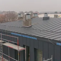 Nowoczesny budynek z ciemną blachodachówką i kominami, widoczny z rusztowania, w pochmurny dzień.
