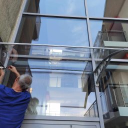 Tech Serwis Dom i Ogród - Montaż zadaszenia nad wejściem do klatki schodowej: pracownik dokręca śruby mocujące konstrukcję z metalowym wspornikiem i przezroczystym panelem, widoczne okna i drzwi wejściowe pod zadaszeniem.