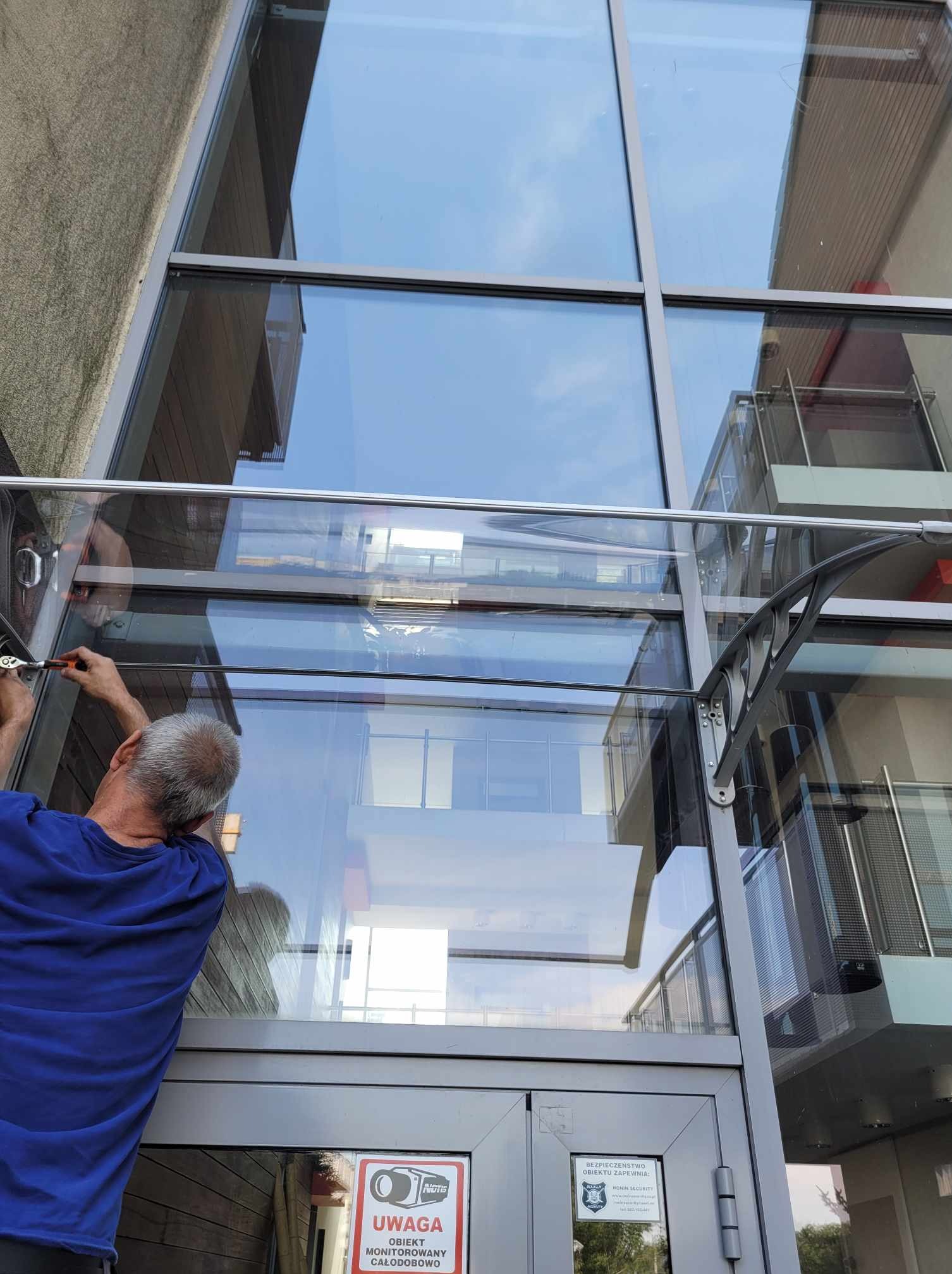Montaż zadaszenia nad wejściem do klatki schodowej: pracownik dokręca śruby mocujące konstrukcję z metalowym wspornikiem i przezroczystym panelem, widoczne okna i drzwi wejściowe pod zadaszeniem.