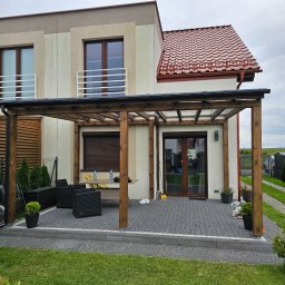 DEM-DREW Damian Mizak - Nowoczesny taras z drewnianą pergolą przy domu z czerwoną dachówką. Na tarasie stoją czarne meble ogrodowe i donice z roślinami. Widoczne okna z roletami.