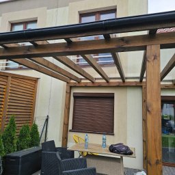 DEM-DREW Damian Mizak - Drewniana pergola z meblami ogrodowymi na tle domu z brązowymi roletami. Widoczne elementy konstrukcyjne i detale wykończeniowe, tworzące przyjemną przestrzeń.