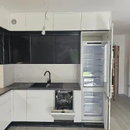 Nowoczesna kuchnia z czarnymi frontami szafek i białymi górnymi szafkami. Widoczny zlew, bateria, marmurowe płytki i otwarta lodówka. W tle widoczne pomieszczenie.