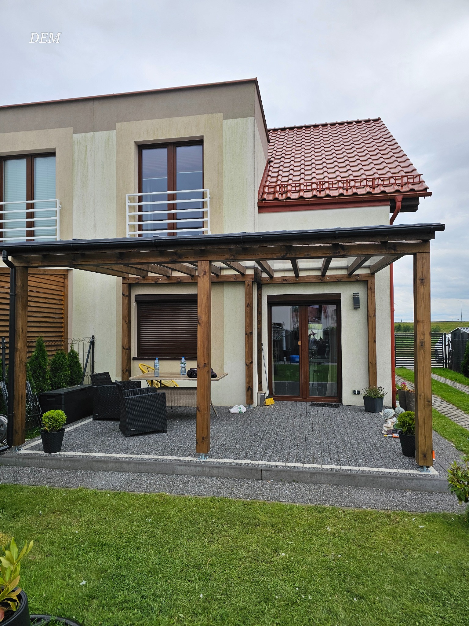 Nowoczesny taras z drewnianą pergolą przy domu z czerwoną dachówką. Na tarasie stoją czarne meble ogrodowe i donice z roślinami. Widoczne okna z roletami.