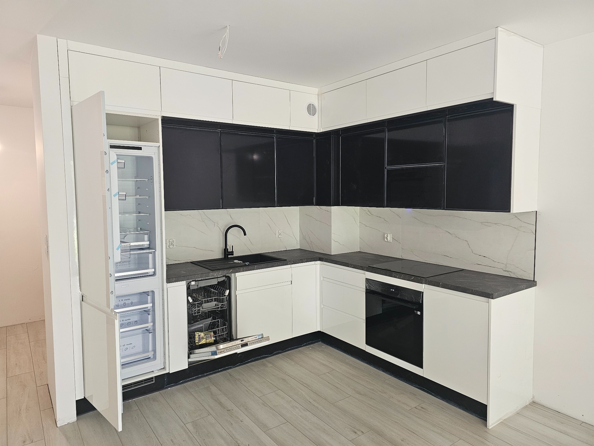 Nowoczesna kuchnia w bieli i czerni z otwartą zmywarką i lodówką w zabudowie. Marmurowy wzór na ścianie, szare panele podłogowe. Minimalistyczny design.