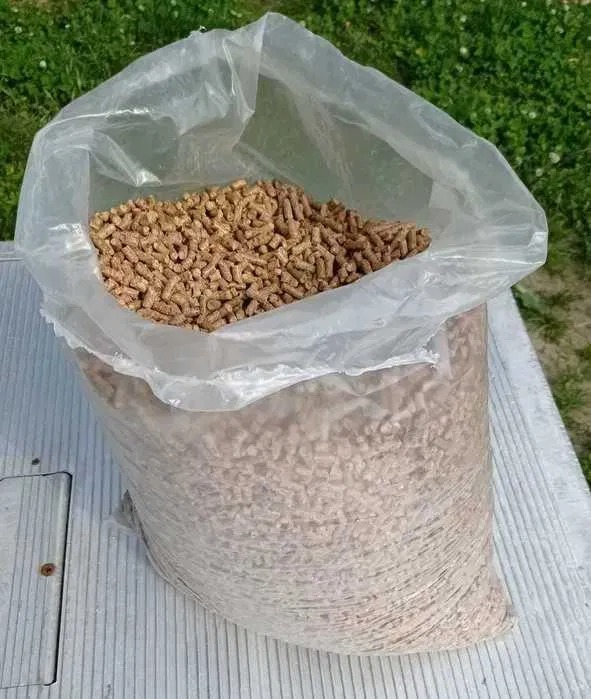 Wypełniony brązowawymi granulkami przezroczysty worek foliowy, częściowo otwarty, leżący na szarej, płaskiej powierzchni na tle trawy.