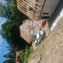 Budowa domu jednorodzinnego w stanie surowym otwartym: ściany z betonu komórkowego, drewniana konstrukcja dachu, biały samochód dostawczy na działce.