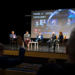 Panel dyskusyjny na scenie z uczestnikami siedzącymi na krzesłach, za nimi ekran z prezentacją i zdjęciami prelegentów, widok z publiczności, osoba podnosi rękę w geście pytania.