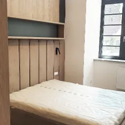 Nowoczesna sypialnia z beżowym łóżkiem z tapicerowanym zagłówkiem, drewnianą zabudową ściany z półkami i okrągłą lampą wiszącą z drewnianych listewek.