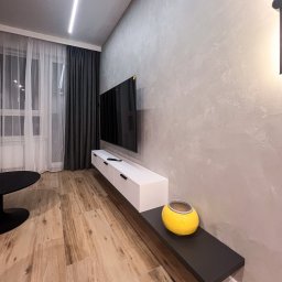 Patrycja Kucharek Studio10 - Nowoczesny salon z podwieszaną białą szafką RTV, telewizorem na ścianie, drewnianą podłogą i szarą ścianą z dekoracyjnym tynkiem.