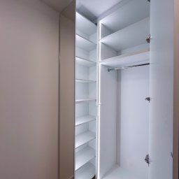 Patrycja Kucharek Studio10 - Wnętrze otwartej, białej szafy wnękowej z oświetleniem LED we Wrocławiu, widoczny system półek i drążek na ubrania, z lustrzanym frontem po lewej stronie.