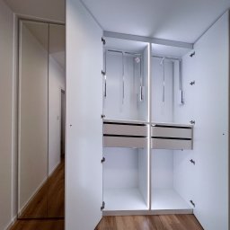 Patrycja Kucharek Studio10 - Wnętrze białej, otwartej szafy z szufladami i drążkami na ubrania, podświetlane pionowymi listwami LED, obok szafa z lustrzanymi drzwiami.