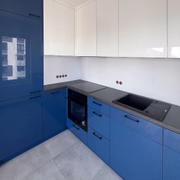 Patrycja Kucharek Studio10 - Nowoczesna kuchnia z niebieskimi, lakierowanymi szafkami dolnymi i białymi górnymi, szary blat, czarny zlew i płyta grzewcza, widoczne niedokończone gniazdka elektryczne na białej ścianie.