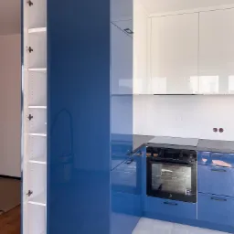 Nowoczesna kuchnia z błyszczącymi, granatowymi szafkami dolnymi i białymi szafkami górnymi, otwarty słupek z półkami po lewej stronie, płyta indukcyjna i piekarnik Electrolux w zabudowie.