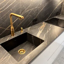 Czarny marmurowy blat kuchenny z złocistymi żyłami, złoty kran i dozownik do mydła, czarny zlew z złotym odpływem, płyta indukcyjna w tle.