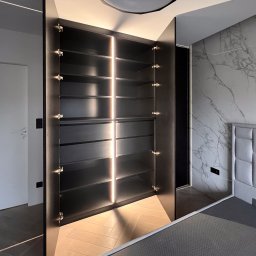 Patrycja Kucharek Studio10 - Otwarta, czarna szafa z podświetlanymi półkami w nowoczesnej sypialni z marmurową ścianą i parkietem w jodełkę.