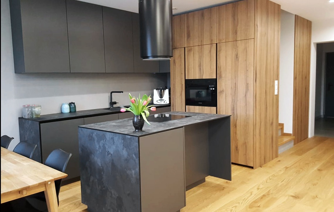 Nowoczesna kuchnia z wyspą, fronty szafek w kolorze grafitowym i drewnopodobnym, czarny okap cylindryczny, blat z efektem kamienia, bukiet tulipanów w wazonie.