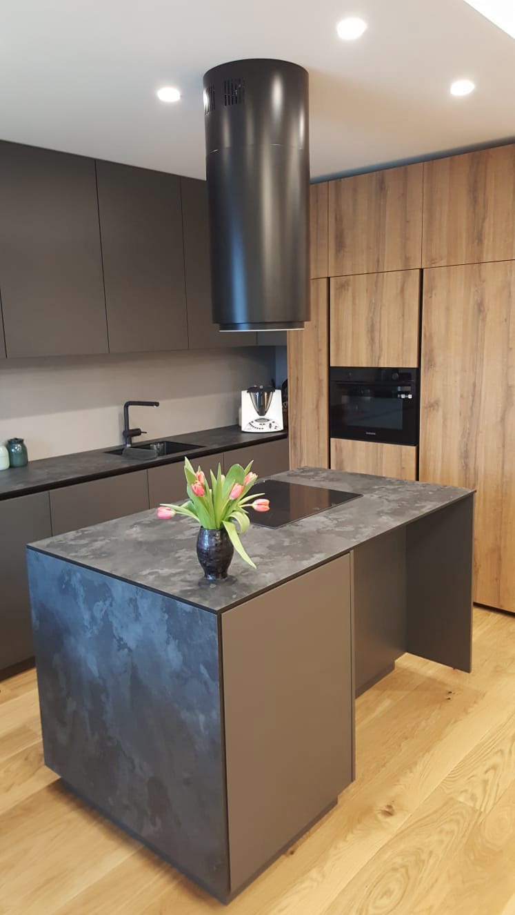 Nowoczesna kuchnia z wyspą, ciemnoszare fronty szafek, drewnopodobne wykończenie wysokiej zabudowy, cylindryczny okap nad wyspą, blat wyspy z efektem kamienia, bukiet tulipanów w wazonie.