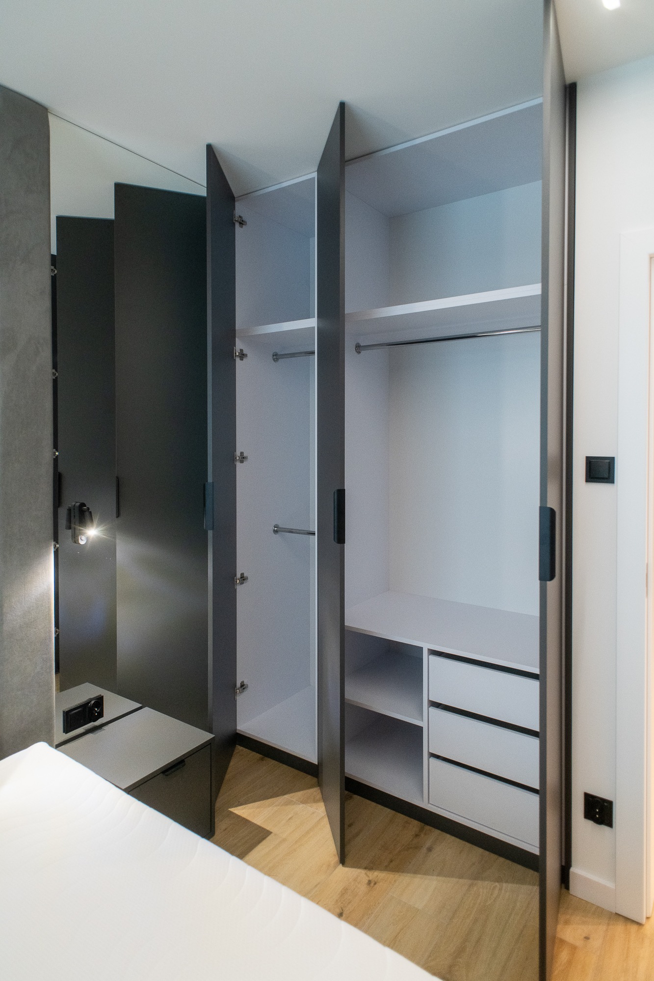 Nowoczesna, otwarta szafa z szarymi drzwiami, białym wnętrzem z półkami, drążkami i trzema szufladami, oświetlona lampką nocną na szafce obok łóżka w sypialni.