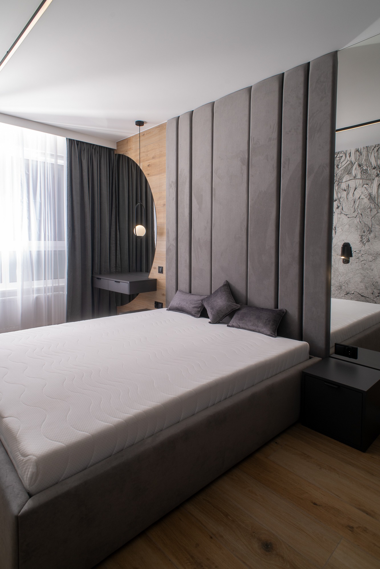 Nowoczesna sypialnia z łóżkiem kontynentalnym, szarym tapicerowanym wezgłowiem w pionowe panele, okrągłą toaletką z lustrem i drewnianym panelem na ścianie, oświetleniem punktowym i zasłonami w oknie.