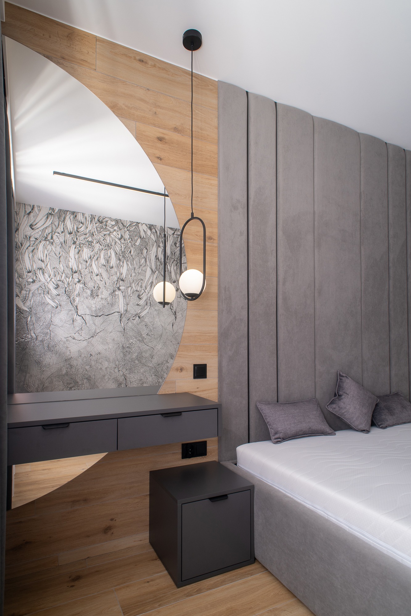 Nowoczesna sypialnia z szarą tapicerowaną ścianą, łóżkiem z szarą ramą i szafkami nocnymi w kolorze grafitowym, toaletka z lustrem o nietypowym kształcie oraz dekoracyjna lampa wisząca z dwoma...