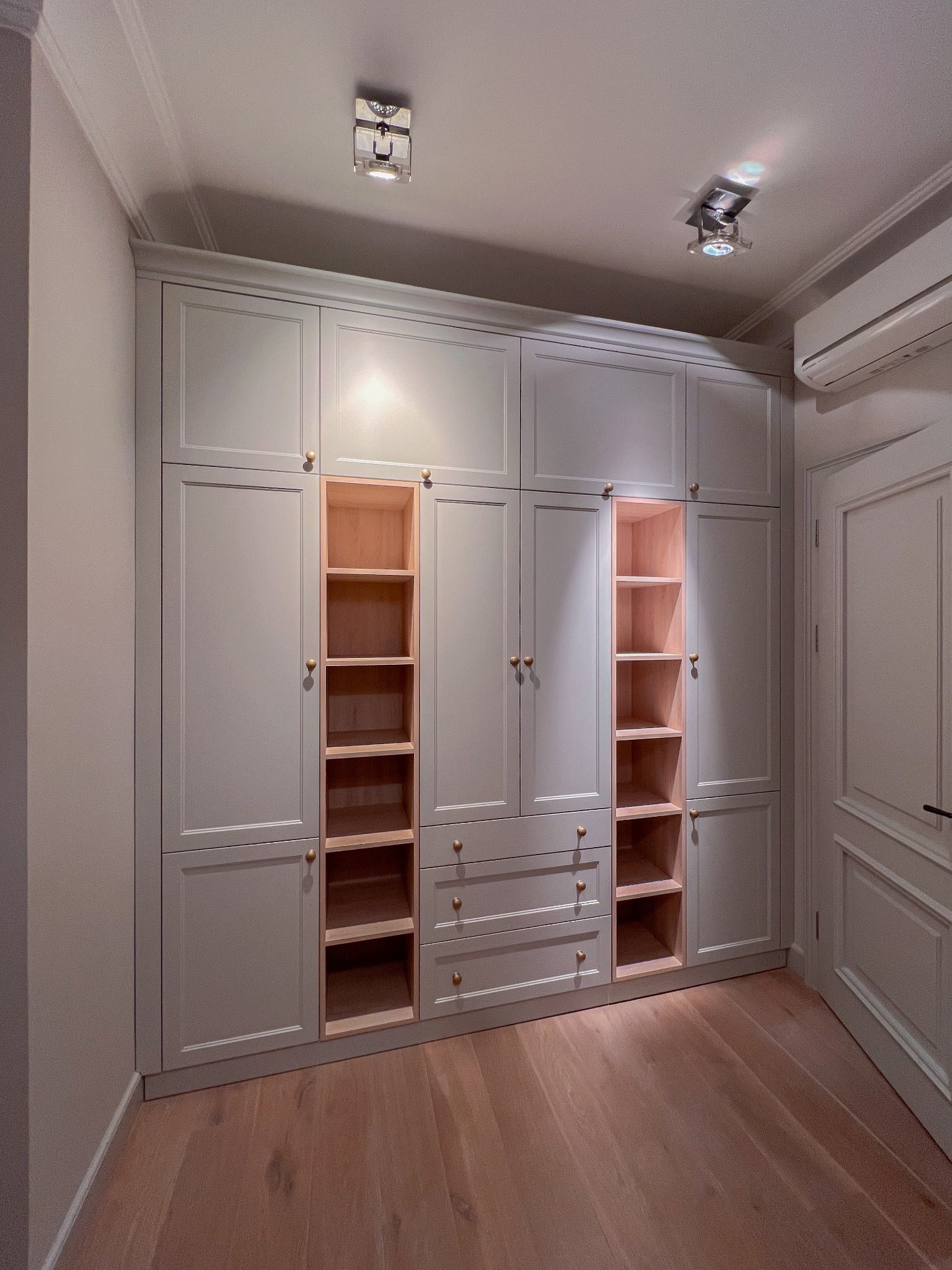 Duża, jasnoszara szafa wnękowa z drewnianymi półkami i złotymi gałkami, wbudowana w ścianę, na podłodze parkiet, oświetlenie punktowe na suficie.