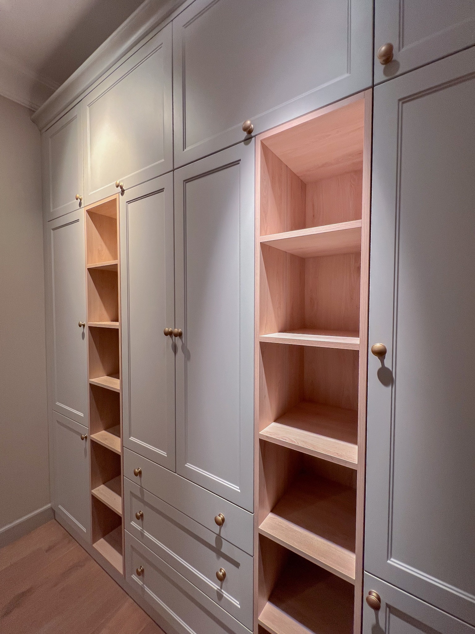 Wielka, szara szafa z drewnianymi półkami i okrągłymi uchwytami, widoczne otwarte półki z surowego drewna bez wykończenia.