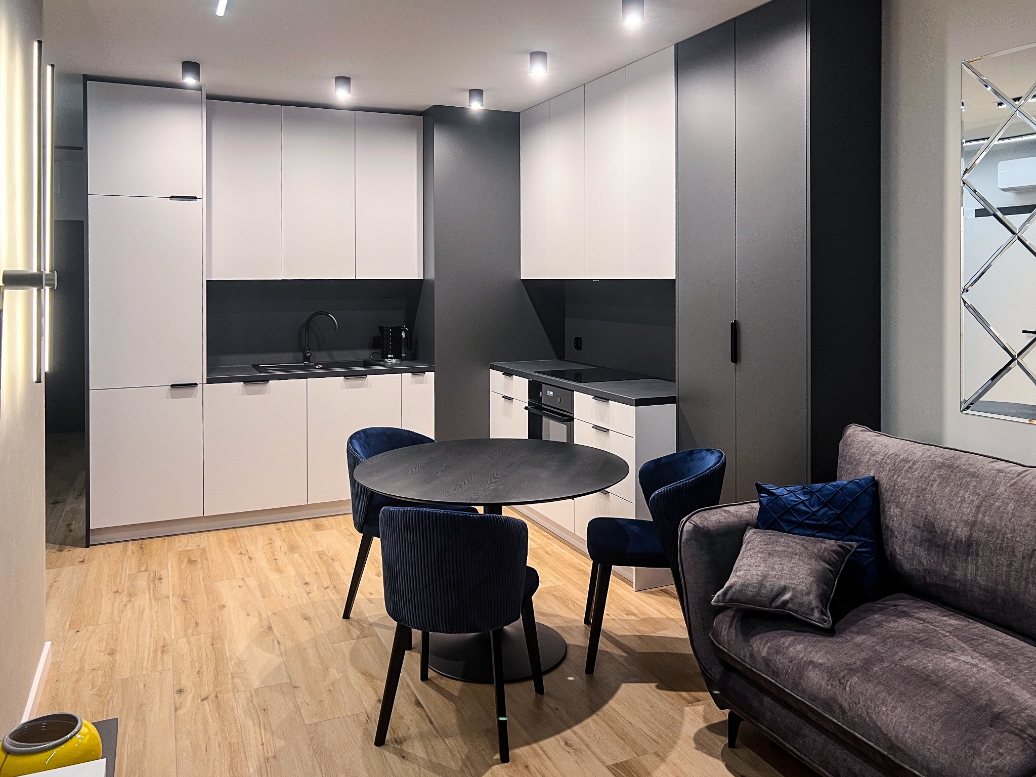 Nowoczesna kuchnia z białymi szafkami, ciemnoszarym blatem, okrągłym stołem z aksamitnymi krzesłami w kolorze granatowym i szarą ścianą, oświetlona punktowymi lampami sufitowymi.