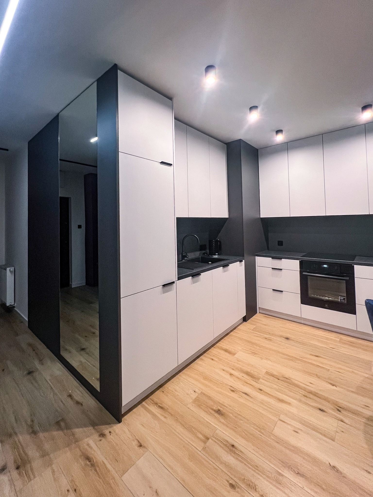Nowoczesna kuchnia z białymi szafkami, czarnymi akcentami i drewnianą podłogą; widoczna zabudowa kuchenna z piekarnikiem i czarną armaturą, obok szafa z lustrzanymi drzwiami.